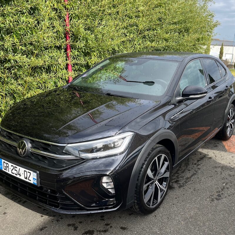 Voiture noire Volkswagen devant haie verte.
